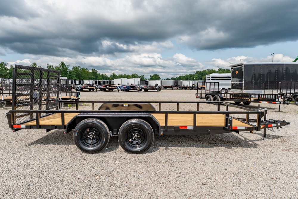 Dual-axle flatbed trailer with a wooden deck and metal rails, parked in a gravel lot with other trailers under a cloudy sky.