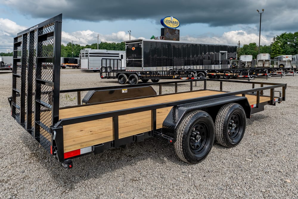 Black flatbed trailer with wooden deck and dual axles on a gravel lot, rear ramp/gate lowered nearby.
