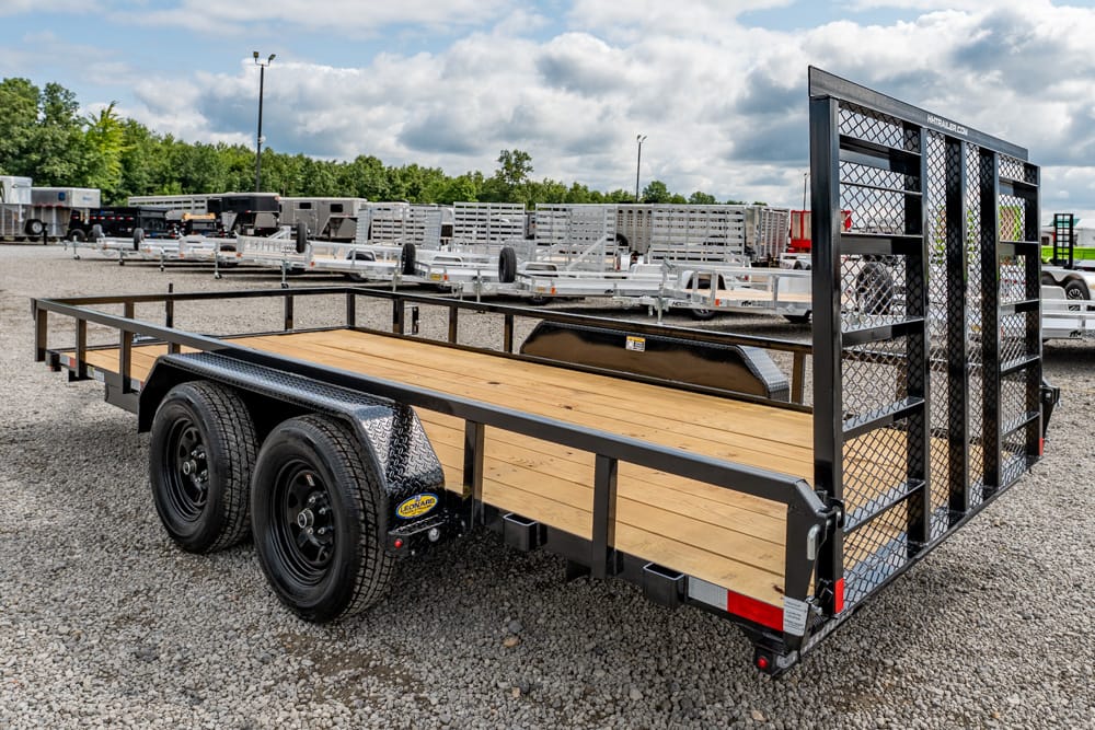 Dual-axle flatbed utility trailer with a wooden deck and tall rear gate on a gravel lot.