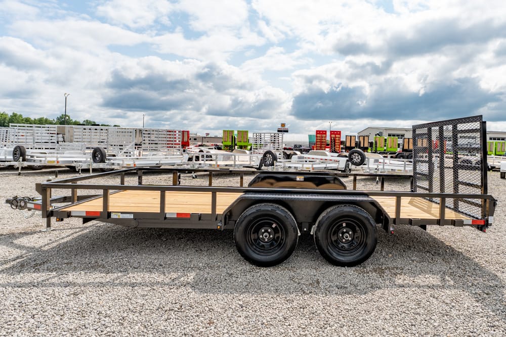 Flatbed utility trailer with dual axles and a rear ramp, parked on gravel lot with other trailers in background