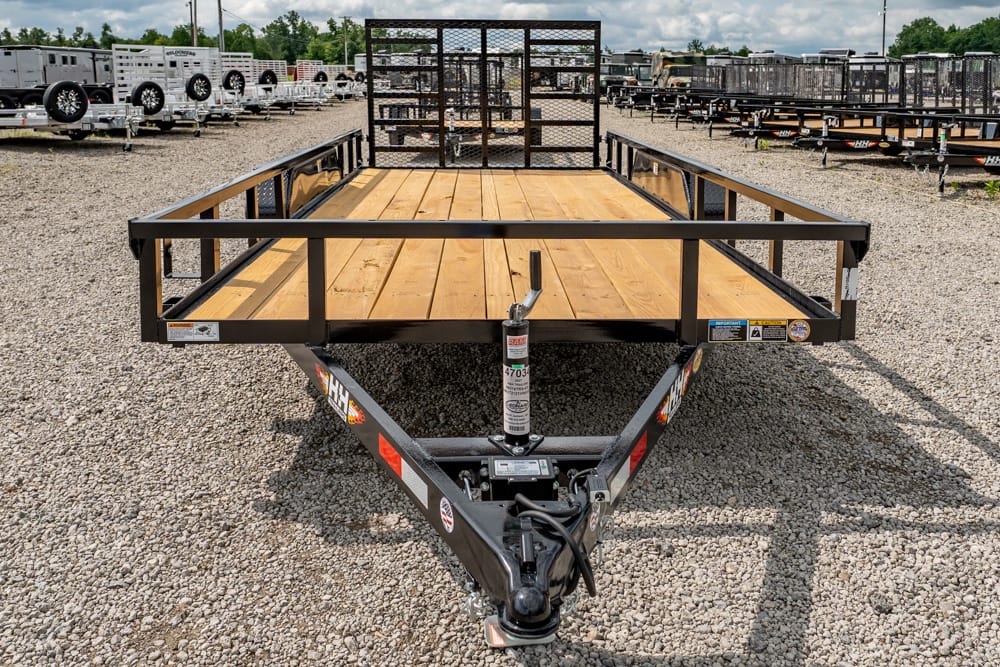 Front view of a flatbed utility trailer with a wooden deck and metal rails on a gravel lot, hitch and jack visible in the foreground.