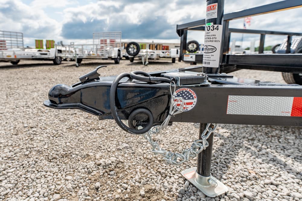 Close-up of a trailer hitch on a gravel lot, showing the coupler, safety chains, and a circular lock with an American flag sticker.