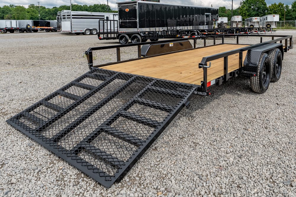 Wide flatbed trailer with a metal ramp folded down, parked on gravel yard, ready for loading or transport of vehicles or equipment