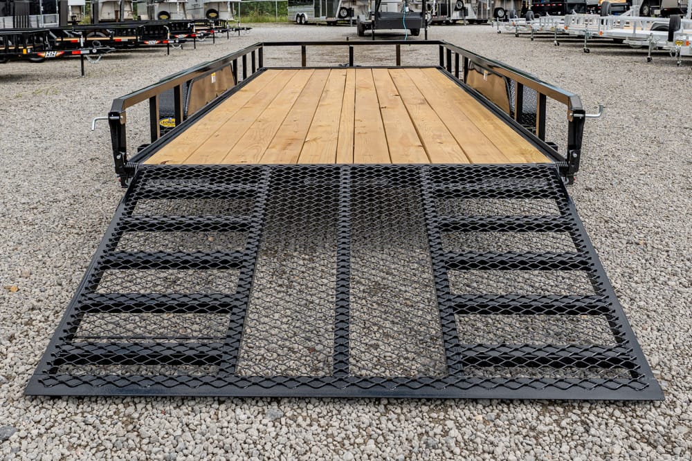 Wide view of a black utility trailer with a wooden bed and a metal mesh ramp extended to the ground on a gravel lot with other trailers in the background.