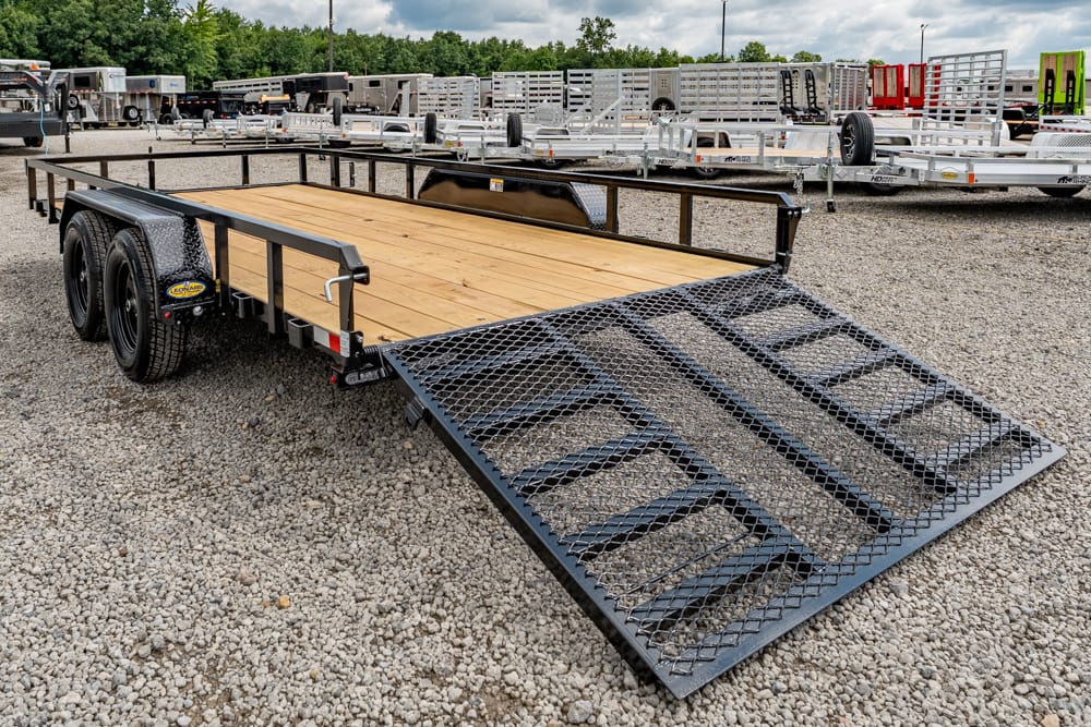 Flatbed trailer with wooden deck and metal ramp extended for loading, parked on a gravel lot with other trailers nearby