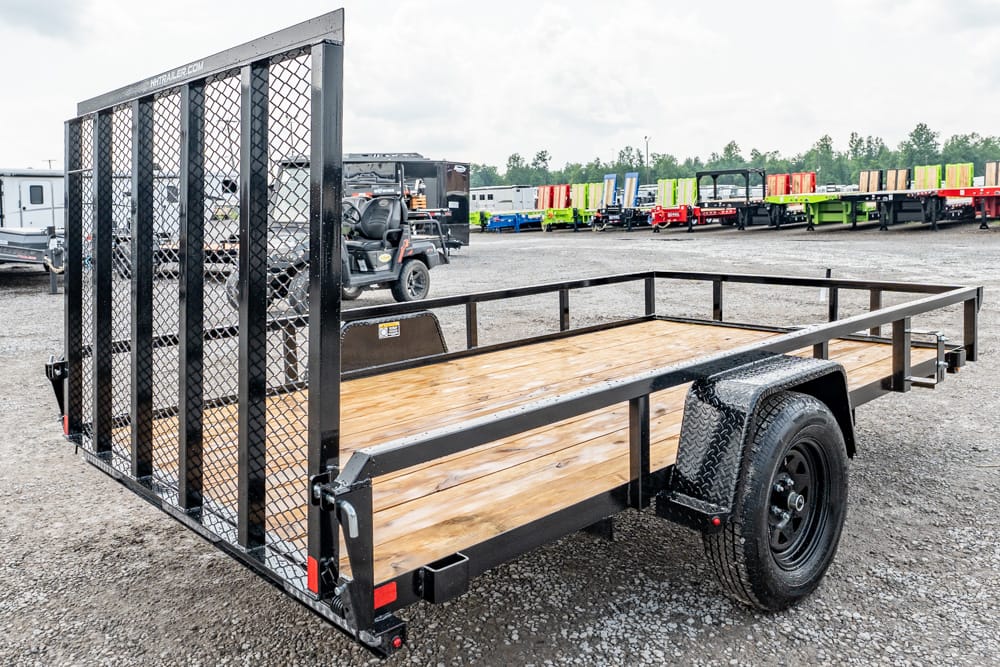 Black metal utility trailer with a wooden deck and mesh ramp, parked on a gravel lot with other trailers in the background.