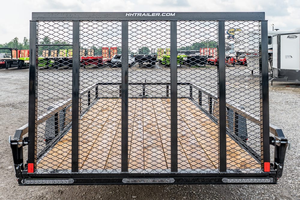 Rear view of a black utility trailer with a metal diamond-mesh gate and wooden deck, parked in a gravel lot with other trailers in the background.