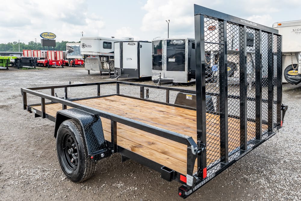 Black utility trailer with a wooden deck and tall mesh rear gate on a gravel lot with other trailers in the background