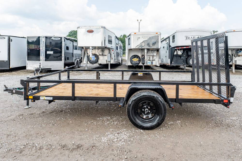 Flatbed utility trailer with a wooden deck and single axle, parked in a lot of enclosed trailers.