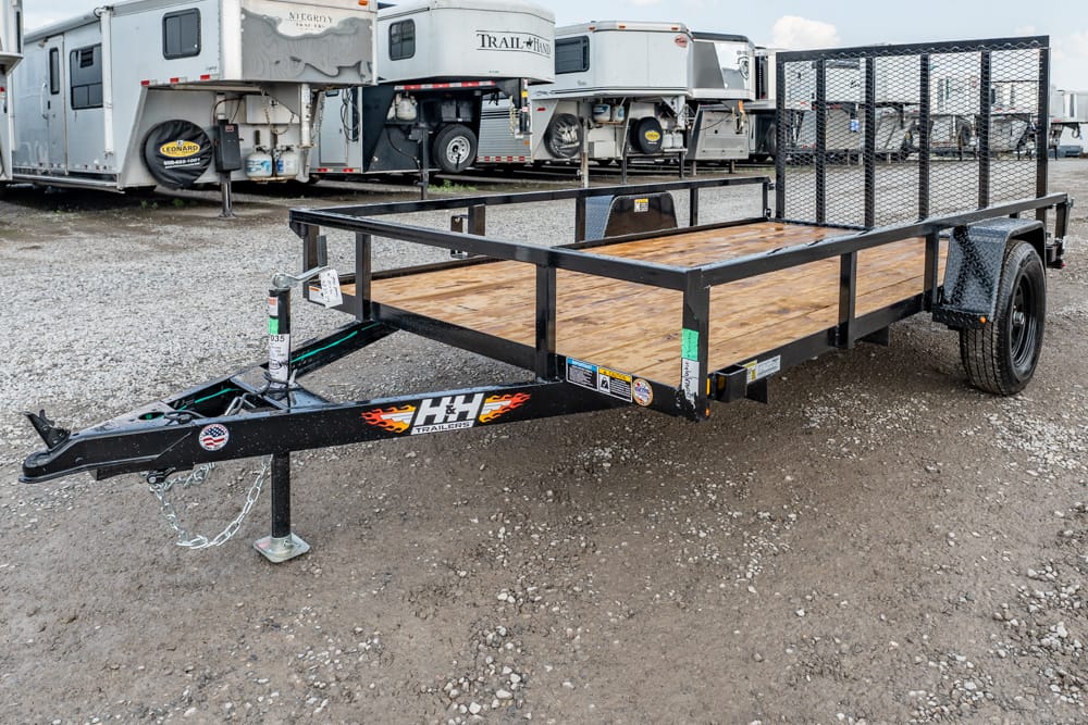 Black flatbed utility trailer with a wooden deck and metal rails, parked on gravel with RV trailers in the background.