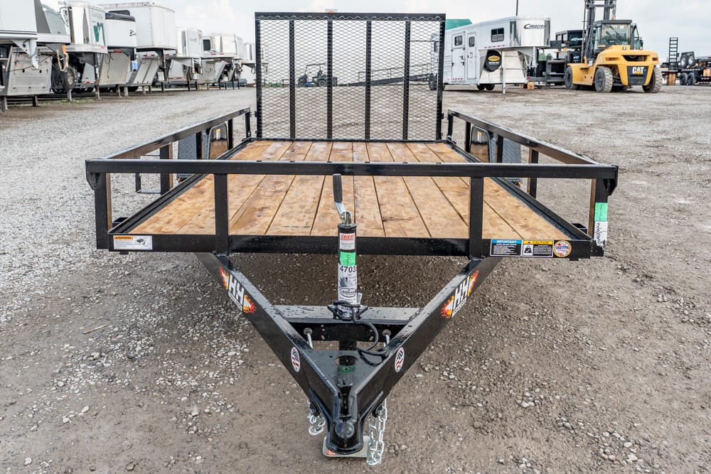 Front view of a black metal utility trailer with a wooden deck and mesh rear gate, parked in a gravel lot.