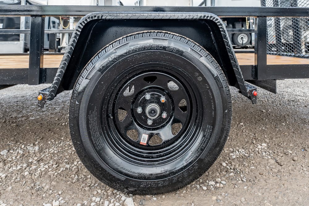 Trailer wheel and tire with a black steel rim, mounted under a metal trailer fender on a gravel surface under a utility trailer.