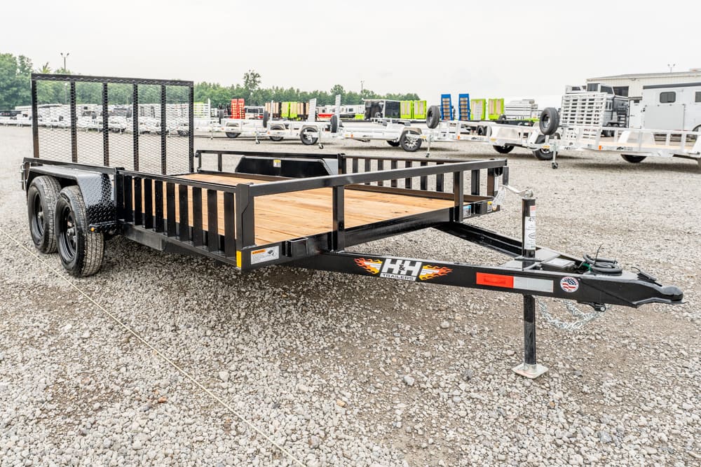 Black flatbed utility trailer with a wooden deck and rear mesh ramp, dual axles, parked on gravel lot.