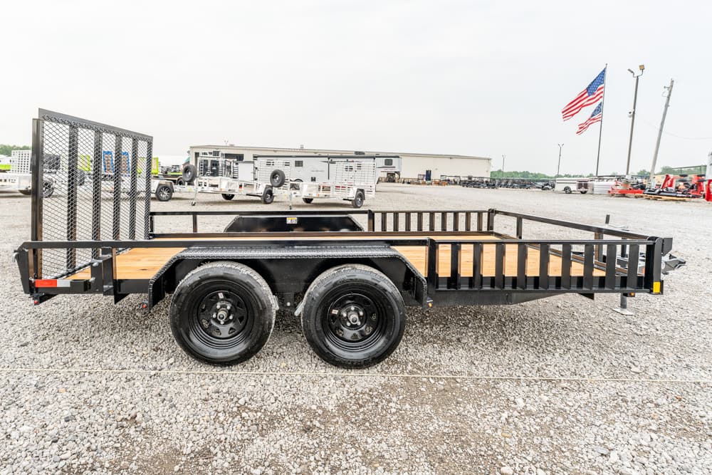 Dual-axle flatbed trailer with a wooden deck and black metal rails on a gravel lot, with an American flag in the background.