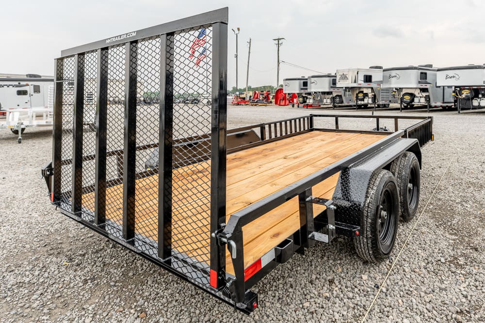 Black dual-axle utility trailer with wooden bed and tall mesh ramp gate on a gravel lot; travel trailers in the background