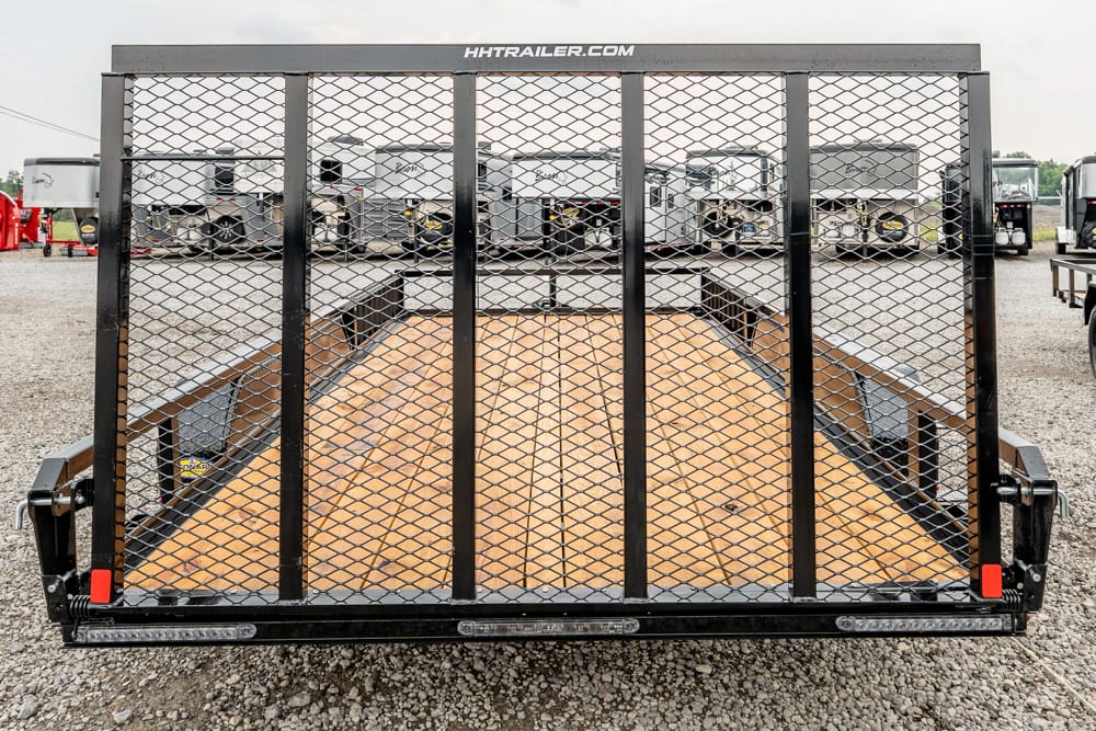 Rear view of a black flatbed trailer with a diamond-mesh gate and wooden deck on a gravel lot, RVs in the background.