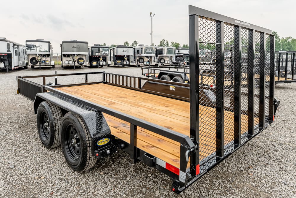 Double-axle utility trailer with a wooden deck and tall mesh ramp, parked on a gravel lot with RVs in the background.