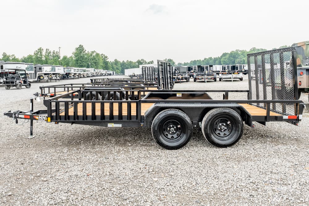Dual-axle flatbed utility trailer with a wooden deck and rear ramp gate, parked on a gravel lot among other trailers.