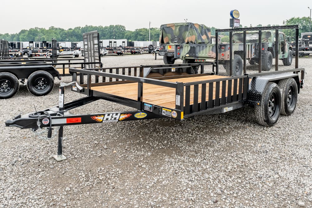 Black dual-axle trailer with a wooden deck and metal railings on a gravel lot; hitch and front jack visible, carry-all trailer with stakes and a mesh rear gate, background shows other trailers.