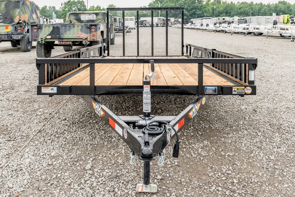 Front view of a black flatbed trailer with a wooden deck on a gravel lot, hitch and jack in the foreground.