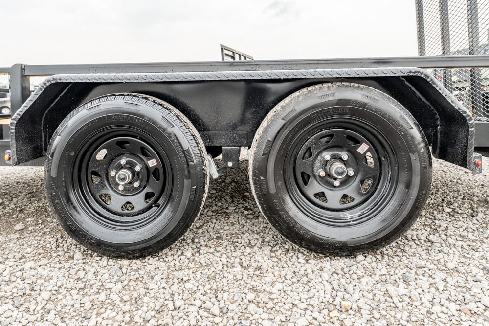 Tandem trailer wheels on a gravel surface with black metal fenders and a utility trailer frame above the tires
