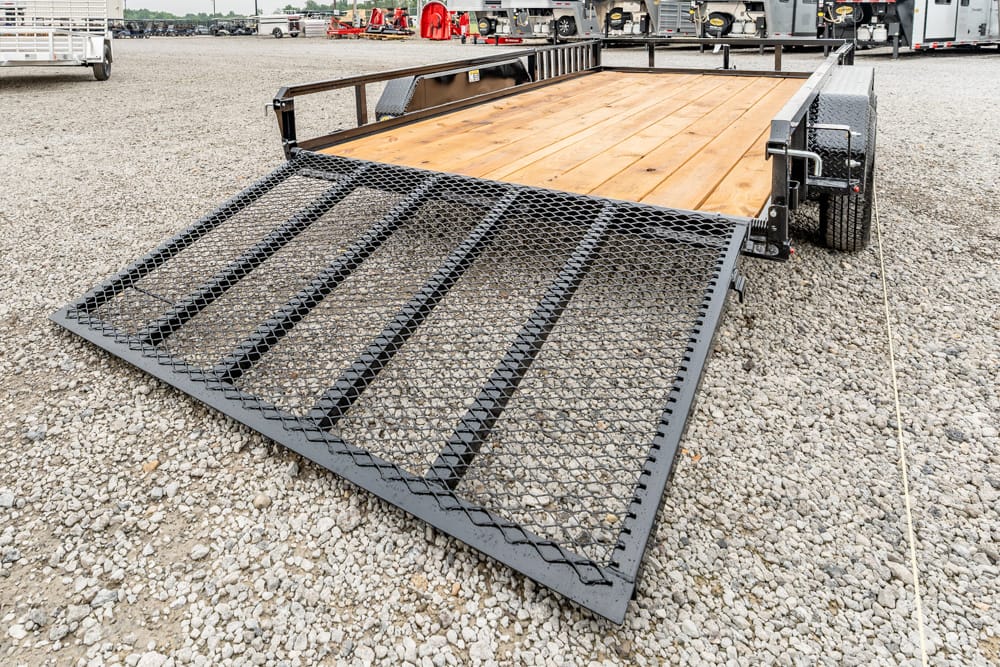 Flatbed trailer with a wooden deck and a lowered metal mesh ramp on a gravel lot, ready for loading