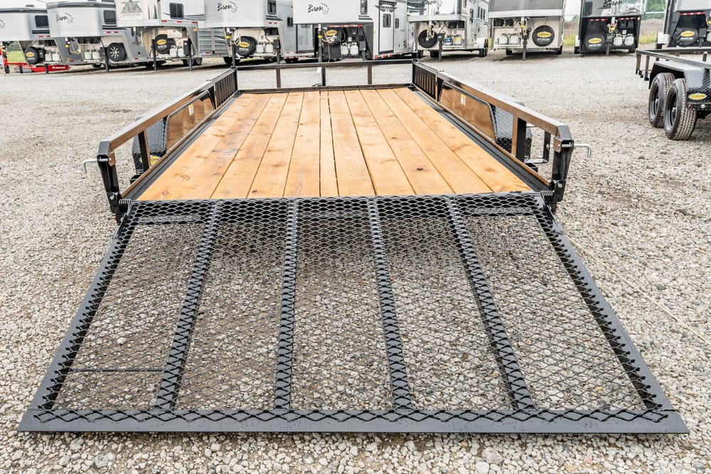 Flatbed utility trailer with a wooden deck seen from the front on a gravel lot, with a diamond-mate ramp at the foreground and side rails visible.