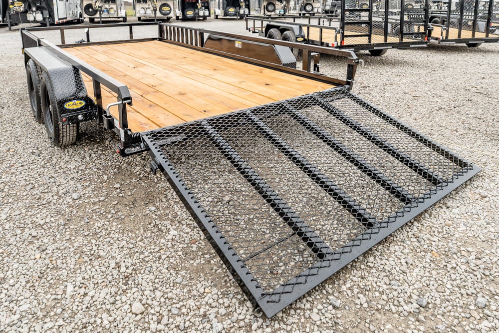 Flatbed utility trailer with a wooden deck and a folded metal mesh ramp, parked on gravel lot in a trailer yard.
