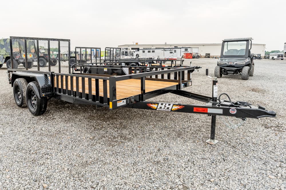 Black utility trailer with wooden deck on a gravel lot, dual axles, and side rails awaiting transport equipment repairs or sales