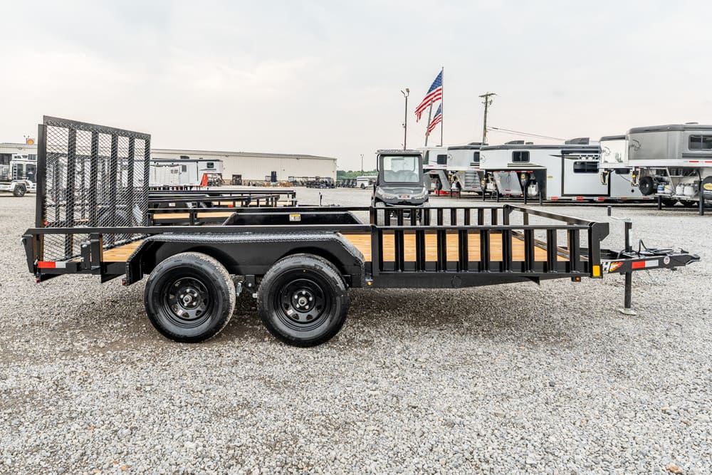 Black flatbed utility trailer with a wooden deck and side rails, dual rear wheels, parked on gravel in a lot with RVs in the background.