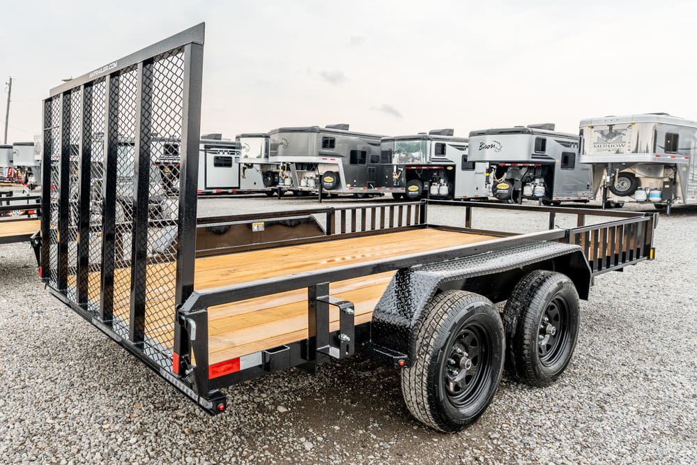 Dual-axle utility trailer with a wooden deck and mesh ramp gate in a gravel lot with RVs in the background.