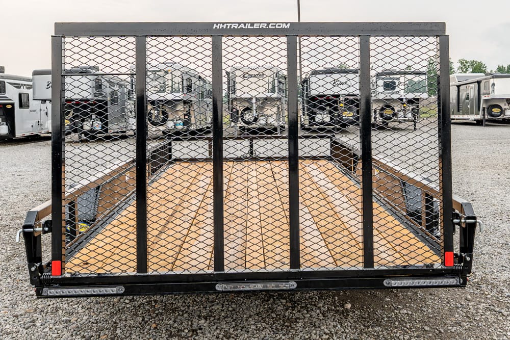 Rear view of a black utility trailer with a mesh partition and wooden deck, parked among other trailers on a gravel lot.