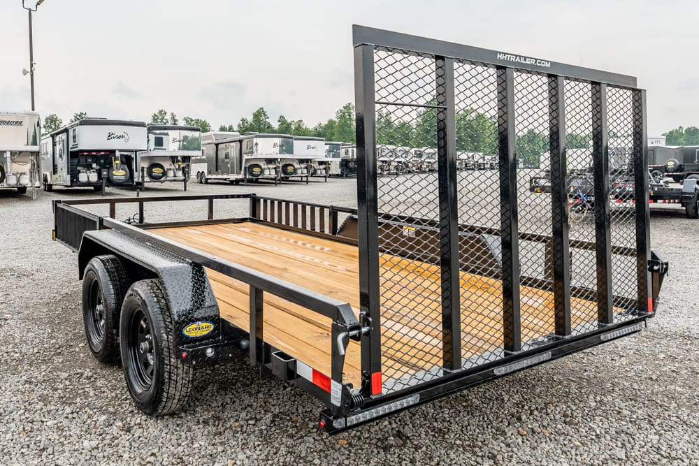 Dual-axle utility trailer with a wooden deck and a tall metal mesh ramp gate, parked in a gravel lot among numerous trailers.