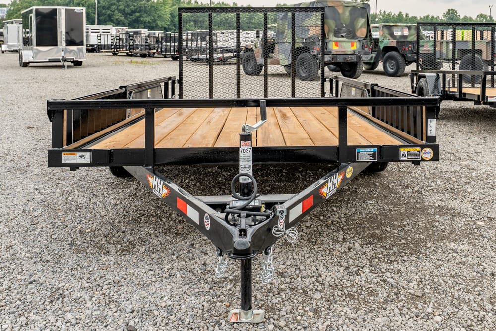 Flatbed trailer with wooden planks in a gravel lot, hitch and safety chains visible, other trailers and military trucks in the background.