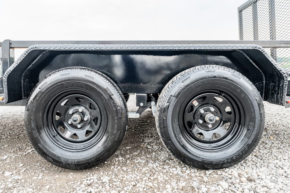 Twin-axle trailer wheels on a black metal trailer, parked on gravel surface.