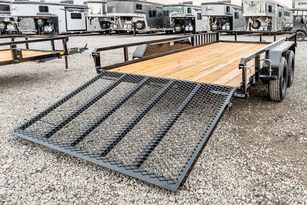 Flatbed trailer with a lowered steel ramp and wooden deck, parked in a lot with other trailers in the background.
