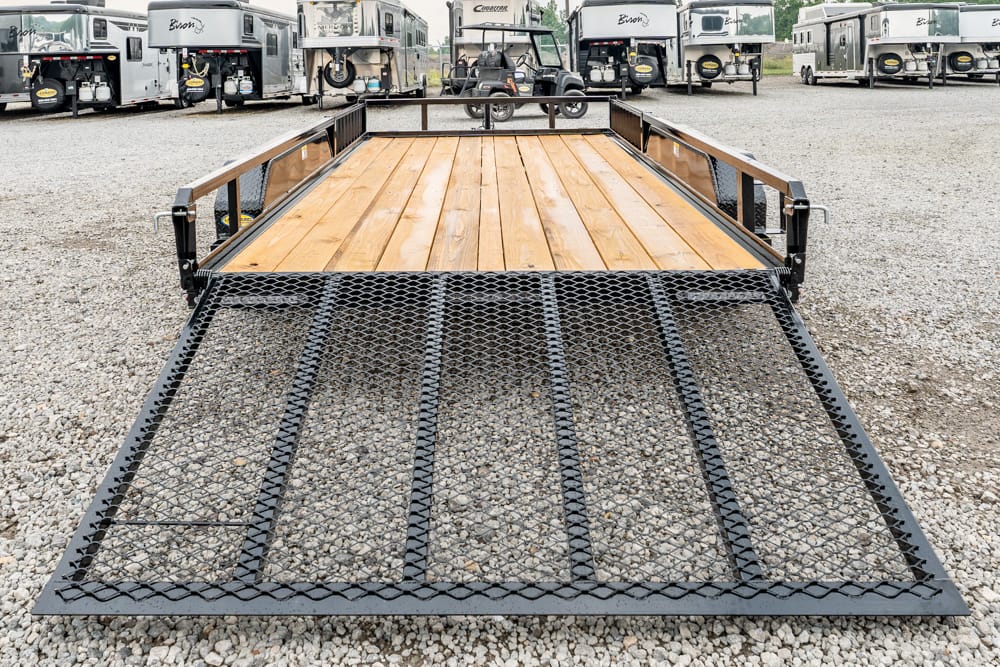 Flatbed trailer with a wooden deck and a perforated steel ramp extended onto a gravel lot, with RVs in the background.