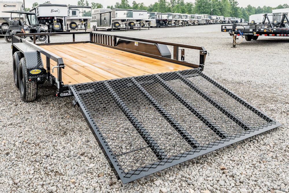 Dual-axle flatbed trailer with a lowered metal mesh ramp on a gravel lot, rows of trailers in the background.