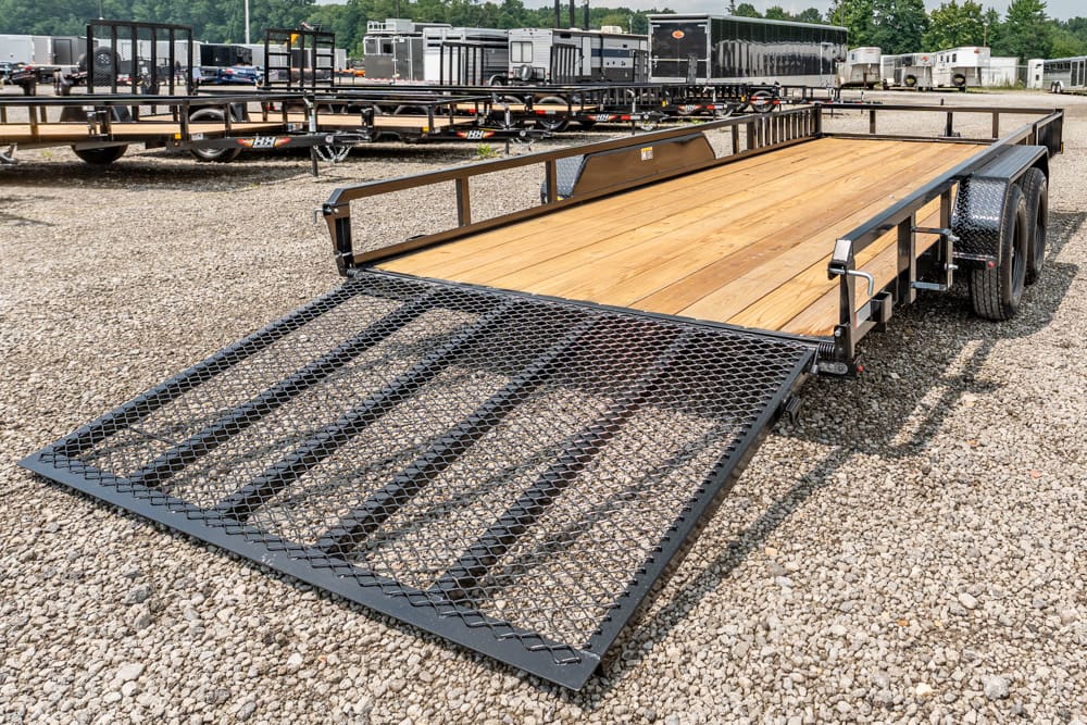 Dual-axle flatbed trailer with wood deck and a down ramp on a gravel lot, ready to load.