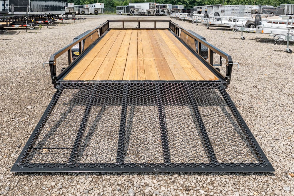 Flatbed utility trailer with a wooden deck and lower mesh ramp on a gravel lot.