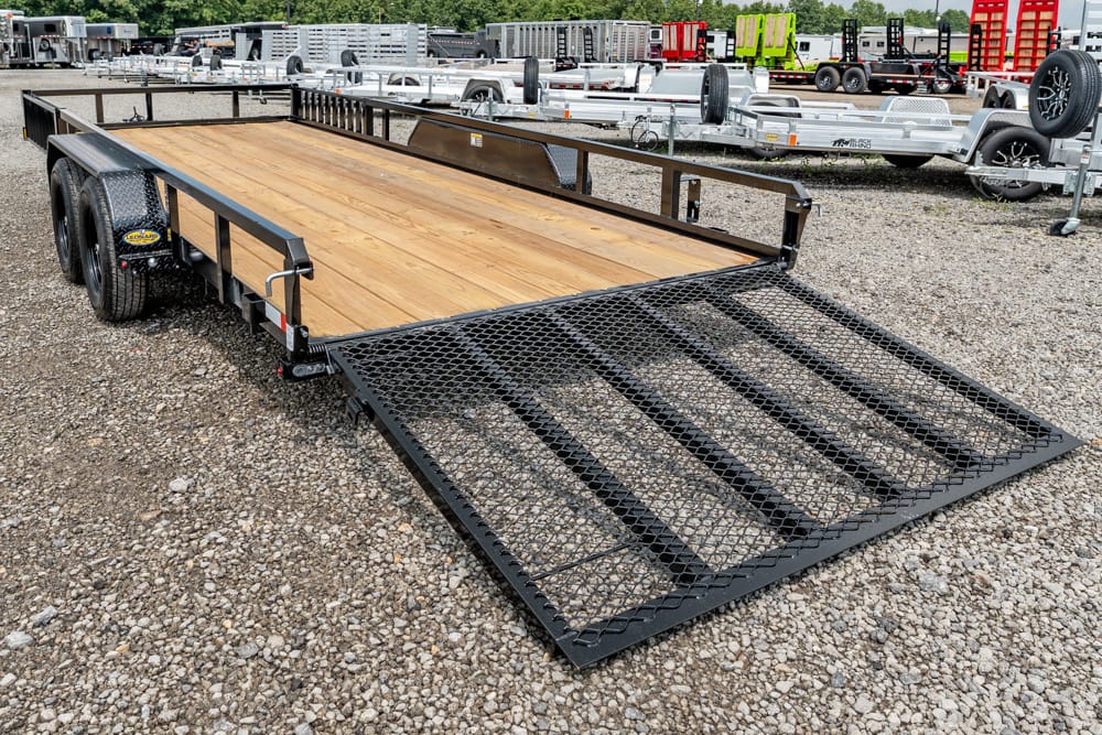 Flatbed trailer with a wooden deck and a fold-down mesh ramp, parked on a gravel lot with other trailers in the background.