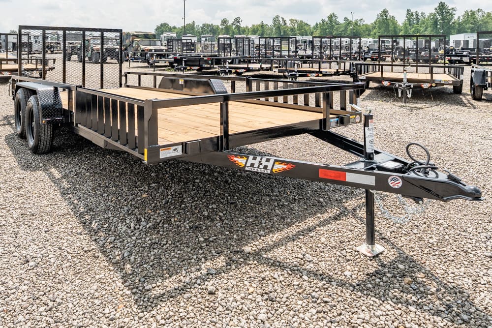 Black steel flatbed trailer with a wooden deck and guard rails, parked on a gravel lot among many other trailers.
