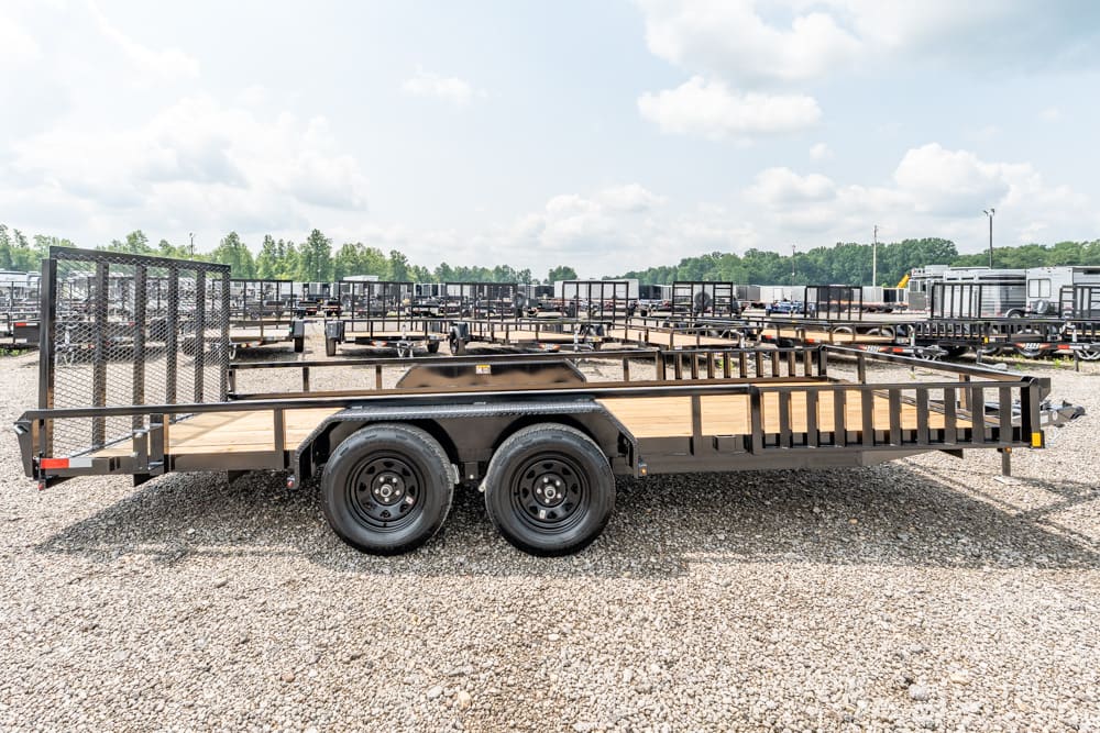 Flatbed utility trailer with dual axles parked in a gravel lot, surrounded by other trailers and equipment.