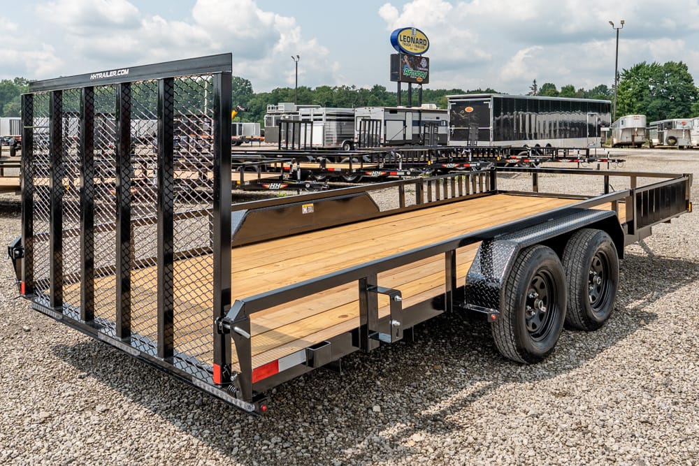 Wide view of a black dual-axle utility trailer with a wooden deck and tall mesh ramp gate in a gravel lot.