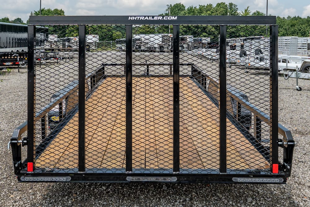 Open flatbed utility trailer with a black metal mesh gate and wooden deck, parked in a gravel lot among other trailers; 'HH TRAILER.COM' is visible on the top rail.