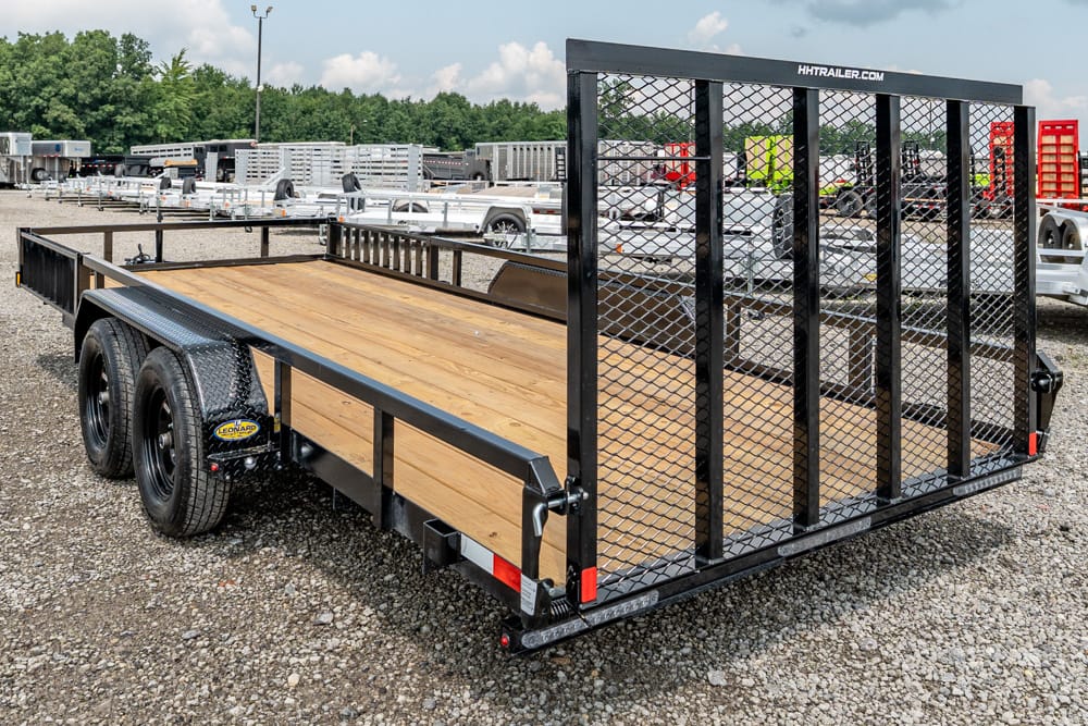 Dual-axle utility trailer with a wooden deck and ramp, parked on gravel in a trailer yard.