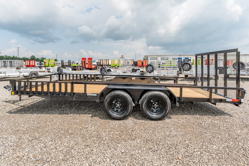 Flatbed trailer with a wooden deck and side rails, shown on a gravel lot with other trailers in the background.