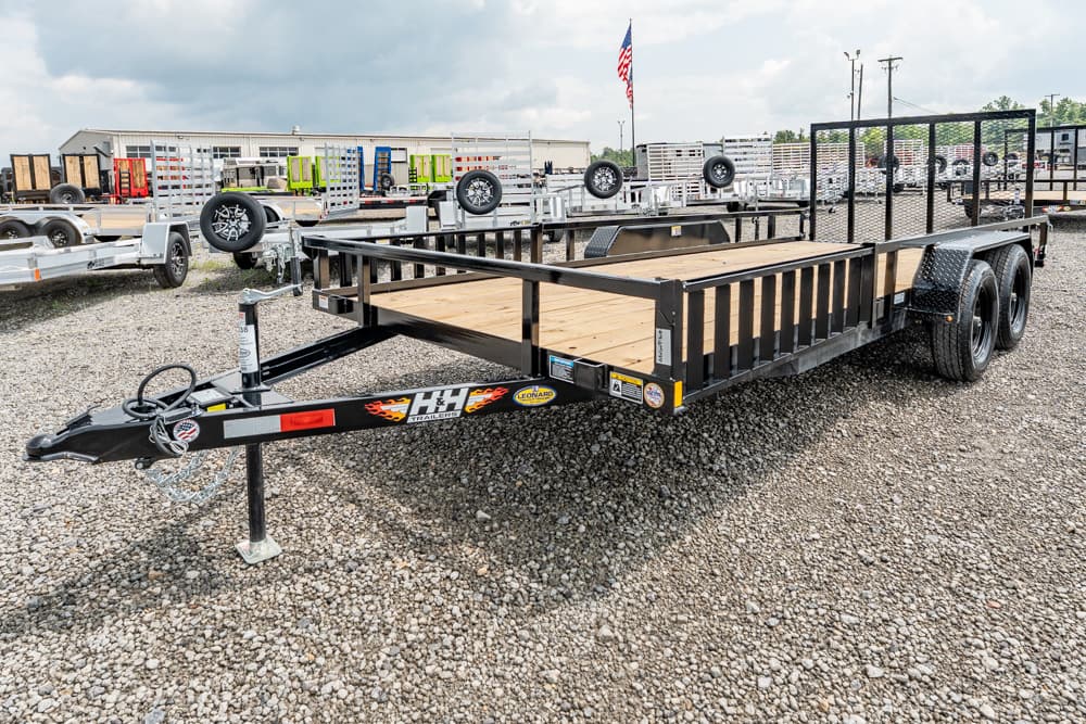 Black dual-axle utility trailer with a wooden deck on a gravel lot, hitch and safety chains in view.