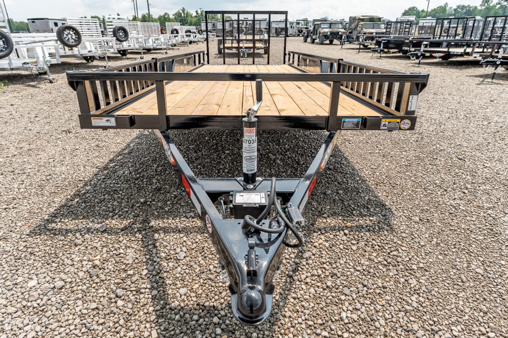 Front view of a flatbed trailer on a gravel lot, black steel frame with a wooden deck and hitch, with a tongue jack and safety chains visible among surrounding trailers.