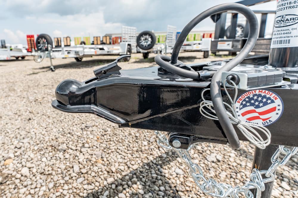 Close-up of a black trailer hitch with safety chains and a circular label on a gravel lot; other trailers are visible in the background.